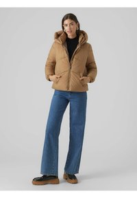Smeđa puff jakna s kapuljačom, a quilted teksturom, s prednjim zatvaračem i dva bočna džepa. Uparena s plavim širokim farmerkama i crnim cipelama.
