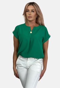 Zielona bluzka z krótkim rękawem i dekoltem w serek, posiadająca dekoracyjny guzik przy dekolcie oraz podwinięte mankiety rękawów, zestawiona z białymi spodniami.