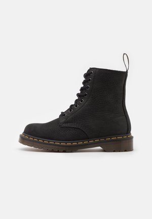 Bottine noire en cuir à lacets avec détail de coutures jaunes, semelle épaisse à rainures, et languette à l'arrière sur fond blanc.