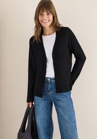 Cardigan nero a maglia con scollatura a V, chiusura frontale con sei bottoni e maniche lunghe, abbinato a jeans blu in denim e una borsa a tracolla nera.