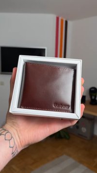 Brown leather wallet in a white box, featuring smooth texture, visible stitching, and embossed "Barbour" logo on the front.