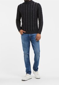 Pull gris foncé en maille torsadée avec un col montant, associé à un jean bleu clair et des baskets blanches, créant un look décontracté.