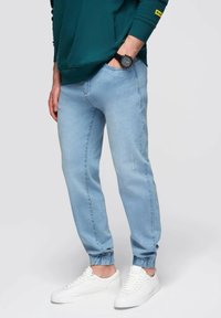 Pantalones de chándal ajustados en azul claro con puños elásticos, hechos de una suave mezcla de algodón. Combinados con zapatillas blancas, con sutiles detalles desgastados visibles.
