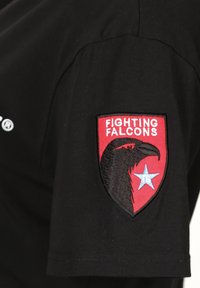 Mustapaita, jossa on punainen brodeerattu tägi hihassa, jossa on falcon-kuvio, teksti "FIGHTING FALCONS" ja sininen tähti. 