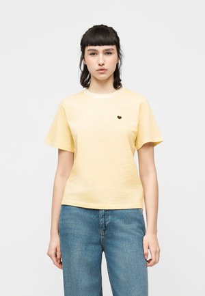 Fiatal nő sötét hajjal, világos sárga, csíkos rövid ujjú felsőt visel kis szív motívummal, valamint kék farmerben, fehér háttér előtt.