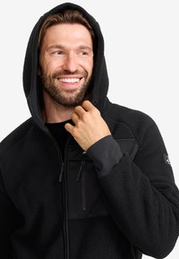 Svart fleecehuva med texturerat tyg, zipfickan på bröstet, justerbar huva och kontrasterande svarta paneler på ärmarna. Släta muddar.