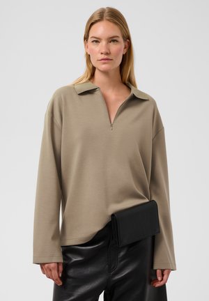 Femme blonde portant un haut beige à manches longues avec un col zippé et un pantalon en cuir noir, tenant une pochette noire à la taille.