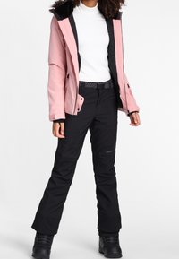Frau in einer pinkfarbenen Winterjacke mit Kapuze, weißem Oberteil, schwarzen Skihosen und schwarzen Schneestiefeln, die vor einem weißen Hintergrund steht.