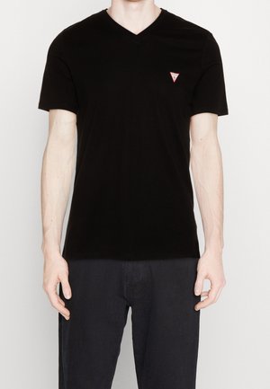 Mężczyzna ubrany w czarny, prosty T-shirt z krótkim rękawem i dekoltem w serek, z małym czerwonym trójkątnym logo na lewej piersi, w połączeniu z ciemnymi spodniami.