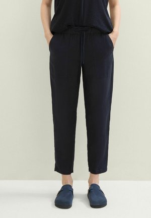 Persoon draagt marineblauwe broek met trekkoord en zakken, en marineblauwe instapschoenen, staand tegen een effen lichte achtergrond.