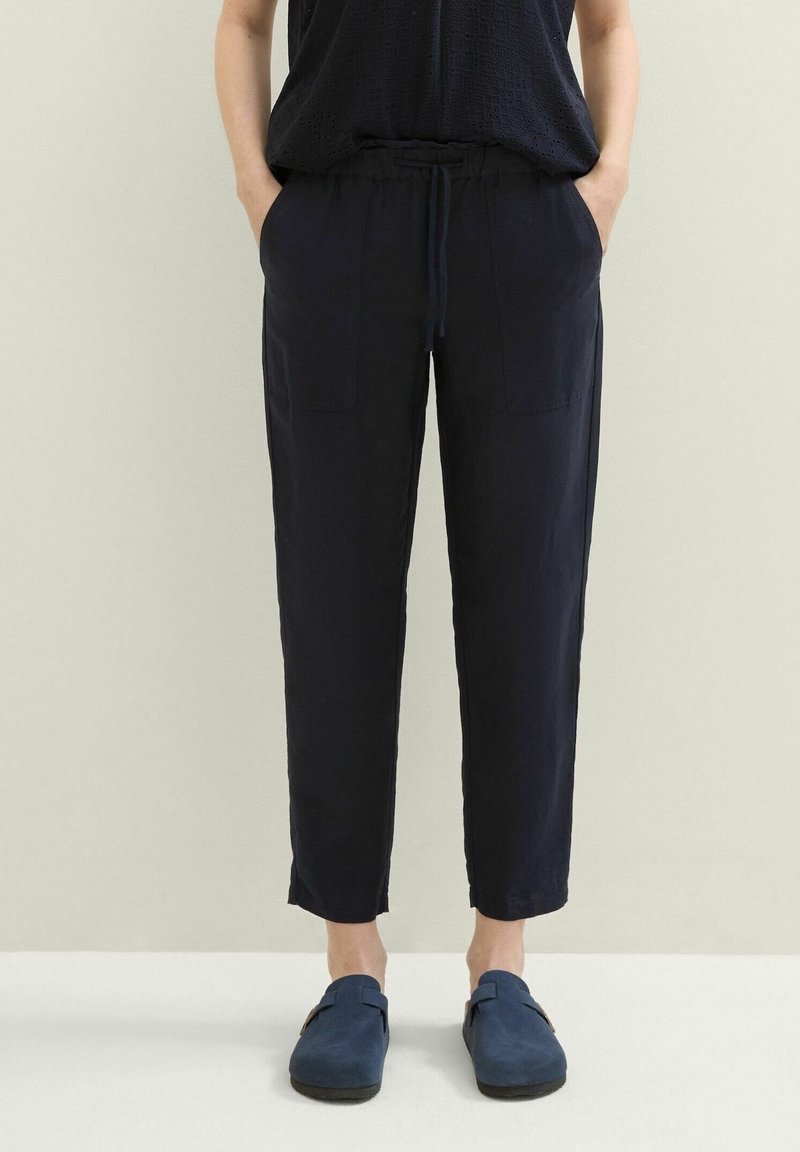 Personne portant un pantalon à cordon de serrage bleu marine avec des poches et des chaussures sans lacets bleu marine, debout devant un fond clair uni.