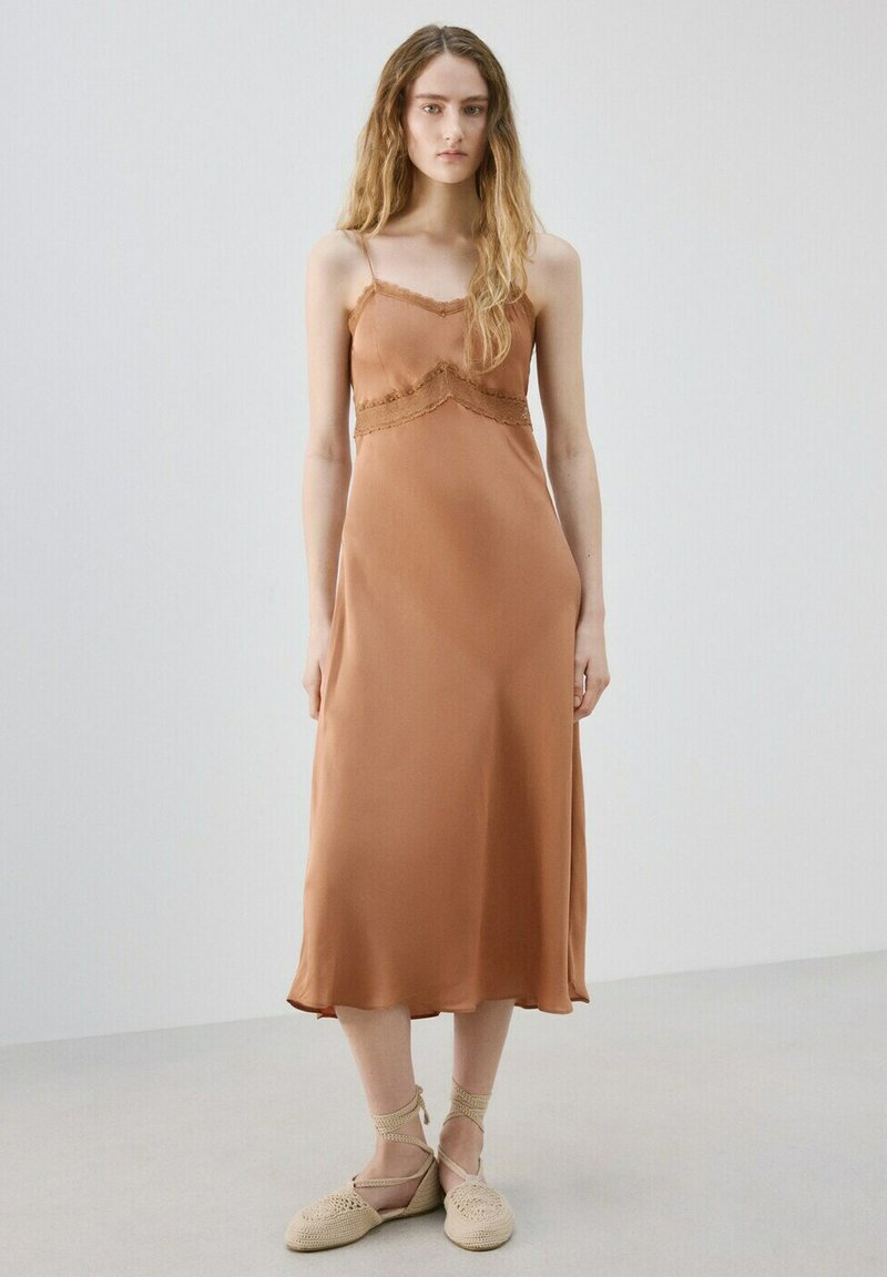 Femme aux longs cheveux portant une robe nuisette midi sans manches couleur cuivre et des chaussures plates en crochet beige, debout contre un mur uni.