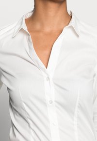 Woman wearing a fitted white button-up shirt with the top two buttons undone against a plain background.