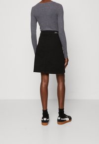 Henkilö, joka on pukeutunut mustiin hameeseen, harmaaseen pitkähihaiseen paitaan, mustiin sukkiin ja musta-valkoisiin tennareihin, seisoo vaalean valkoista taustaa vasten.