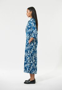 Blaues Blumenkleid mit Dreiviertelärmeln, seitlichen Taschen und lockerer Passform. Der Stoff zeigt weiße und hellblaue Blumenmuster. Schwarze Schuhe.