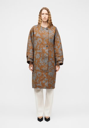 Kobieta w brązowym długim płaszczu z niebieskimi kwiatowymi wzorami, białych spodniach i czarnych spiczastych butach, stojąca na tle jednolitego tła.