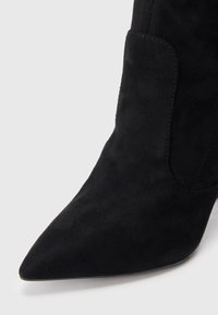 Melnas zamšādas potīšu zābaki ar smailu pirkstu un gludu tekstūru. Iezīmētas ar sānu paneļa dizainu, kas nodrošina elastību, un slaidu, augstu papēdi.