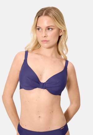 Mujer rubia con bikini azul oscuro con un detalle anudado en la parte superior, mirando ligeramente hacia su izquierda contra un fondo liso.