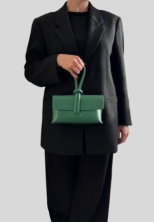 Borsa a tracolla in pelle verde con un design strutturato, caratterizzata da un manico annodato e una chiusura a patta. Tenuta davanti a un blazer nero.