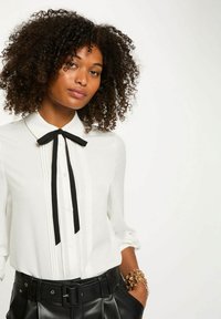 Donna con capelli ricci che indossa una camicetta bianca con fiocco nero e pantaloni di pelle neri con cintura, mano in tasca, su sfondo bianco uniforme.