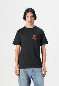 T-shirt de algodão preta com mangas curtas e decote redondo. Apresenta o texto laranja "RIDE THE RAILS" impresso no lado esquerdo do peito.