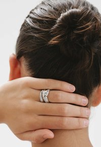 Anillo trenzado de plata con una gema central, llevado en un dedo, mano reposando sobre un cuello. Cabello recogido en un moño, mostrando piel y oreja.