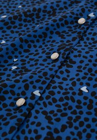Tissu bleu avec des taches noires irrégulières et de petites formes de cœur blanches, avec des boutons-pression argentés le long d'une couture.
