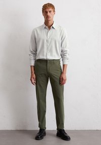 Jasnoszara koszula z długim rękawem zapinana na guziki, zestawiona z oliwkowymi spodniami. Model nosi czarne buty sznurowane, stojąc na neutralnym tle.