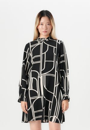 Young woman with straight ombre hair wearing a black and white geometric patterned long-sleeve dress, standing against a plain white background.