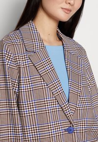Žena na sobě má hnědo-modro-bílou kostkovanou sako přes světle modrý top, na kterém je modrý knoflík vepředu.