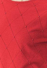 Piros kötött felső, amely gyémántmintával rendelkezik, minden kereszteződésnél apró kerek gyöngyökkel díszítve, sima textúrával.