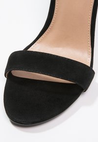 Sandale à talon haut en daim noir à bout ouvert avec une seule bride avant et un détail de couture sur la semelle intérieure beige rembourrée.