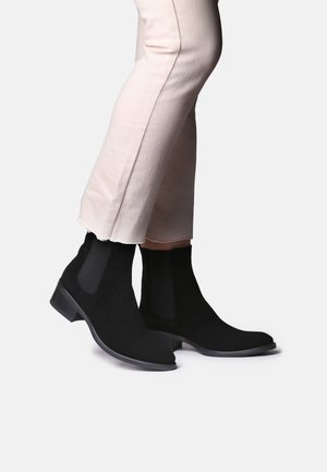 Juodos suede Chelsea botai su elastingais įdėklais, su apvaliu padu ir žemais blokiniais kulniukais, derinami su šviesiai smėlinėmis kelnėmis.