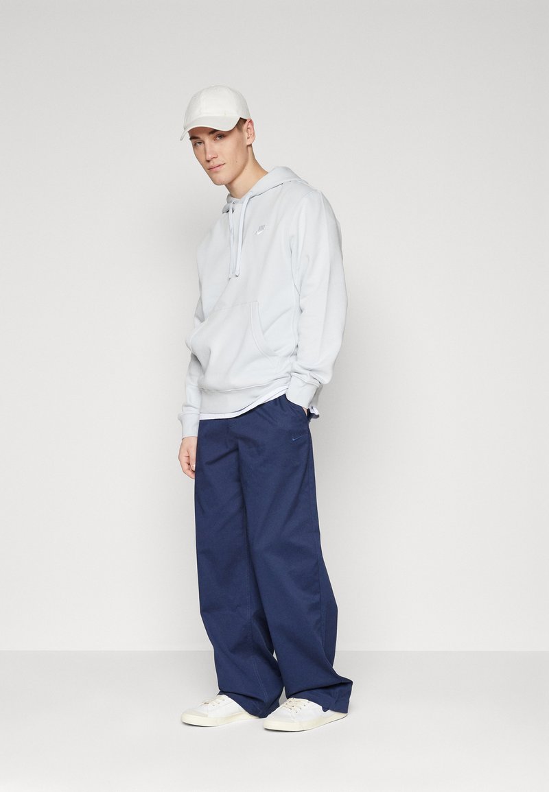 Felpa azzurra chiara con tasca frontale e logo bianco, abbinata a pantaloni navy larghi e sneakers bianche. Design semplice e texture morbida.