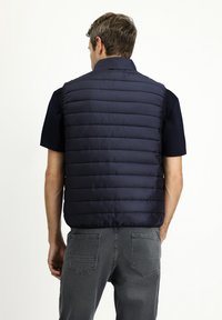 Granatowa kamizelka puchowa o pikowanej strukturze, z wysokim kołnierzem i przeszyciami, połączona z czarną koszulą z krótkim rękawem oraz szarymi dżinsami.