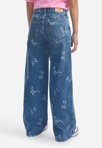 Pogled straga na osobu koja nosi široke plave traperice s bijelim uzorcima mašni i rozu majicu, koja stoji na bijeloj pozadini.