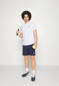 Camiseta blanca de manga corta con el logo de FILA, pantalones cortos azul marino, sosteniendo una raqueta de tenis y una pelota, usando zapatillas con detalles en rojo y blanco.