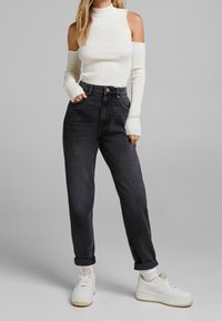 Top bianco a maniche lunghe con spalle scoperte, abbinato a jeans grigio scuro a vita alta e sneakers bianche con suola beige.