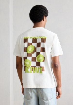 Άνδρας που φοράει λευκή μπλούζα με καφέ καρό μοτίβο, εικόνες πράσινου ακτινιδίου και το κείμενο «MARC O'POLO DENIM KIWI» στην πλάτη.