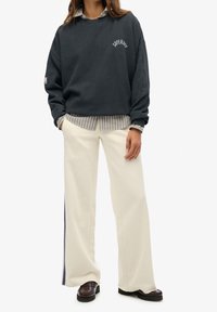 Osoba nosząca granatowy sweatshirt "Superdry" na paskowanej koszuli, kremowe szerokie spodnie z granatowym paskiem oraz ciemno-brązowe loafery.