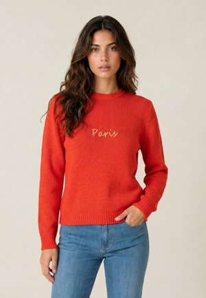 Donna con lunghi capelli scuri che indossa un maglione rosso con il testo "Paris" e jeans blu, in piedi davanti a uno sfondo chiaro e uniforme.