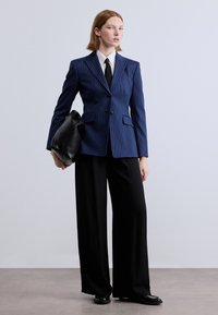 Μπλε ριγέ σακάκι με σκισμένα πέτα, συνδυασμένο με λευκό πουκάμισο και λεπτό μαύρο γραβάτα. Φαρδύ μαύρο παντελόνι και μαύρη τσάντα ολοκληρώνουν την εμφάνιση.