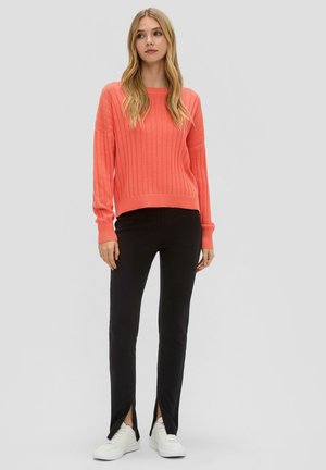 Koraalrood geribbeld sweater met lange mouwen, gecombineerd met zwarte slim-fit broek met zijsplitten, en witte sneakers voor een casual look.