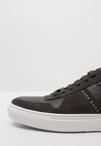 Dunkelgraue Wildledersneaker mit glatten Lederakzenten, weißer Gummisohle, strukturiertem Zehenbereich und auffälliger Markenbeschriftung in schwarzer Schrift.