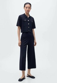 Chemise noire cintrée à manches courtes, avec deux poches poitrine et des boutons dorés, associée à un pantalon noir à jambes larges et des chaussures plates en osier.