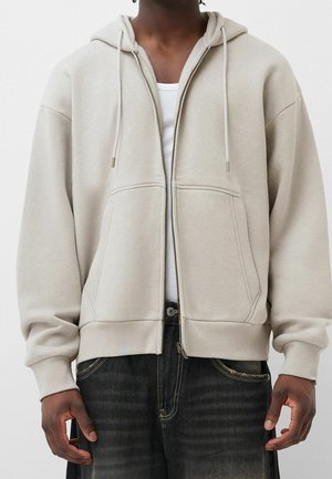 Mand iført beige hættetrøje med lynlås over hvid tanktop og sorte, falmede jeans, stående med hænderne afslappet ned langs siderne.