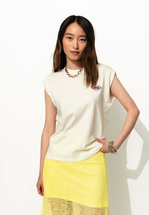 Un t-shirt sans manches crème avec un petit logo bleu et orange, associé à une jupe en dentelle jaune transparente. Les accessoires comprennent un collier massif et des bracelets.
