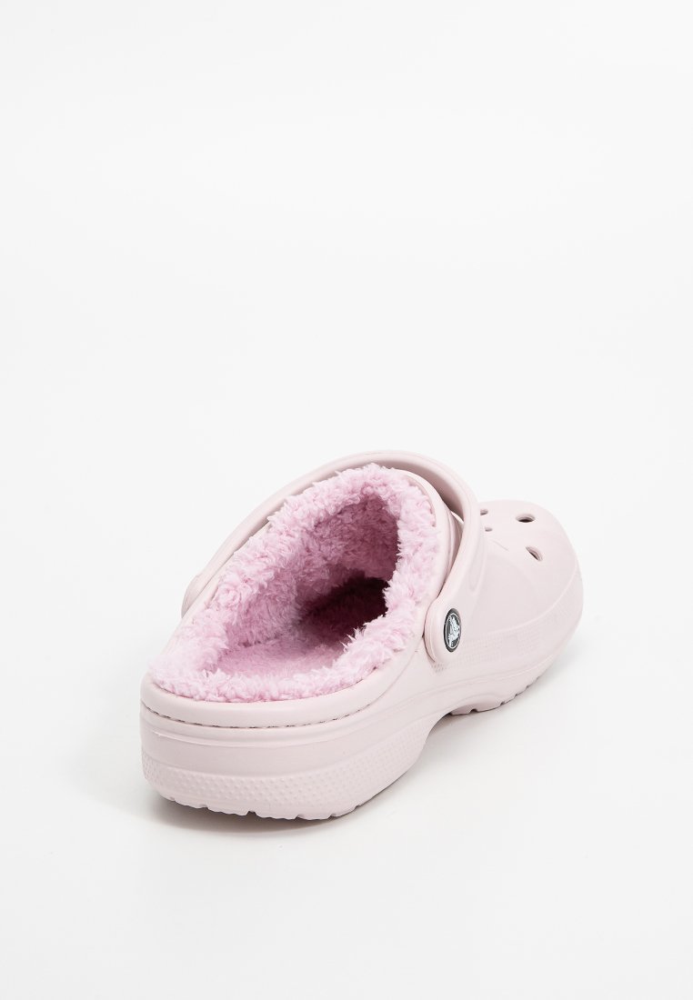 Zueco rosa con un interior suave y esponjoso y orificios de ventilación. Fabricado en material de goma con una suela texturizada y un detalle de logo circular.