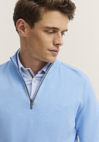 Hellblauer Strickpullover mit einem Viertelreißverschluss-Kragen. Verfügt über ein dezentes, gesticktes Logo auf der Brust und ein gestreiftes Hemd darunter.