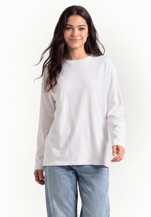 Ung kvinde med langt mørkt hår iført en hvid langærmet skjorte og lyseblå jeans, stående foran en ensfarvet hvid baggrund.